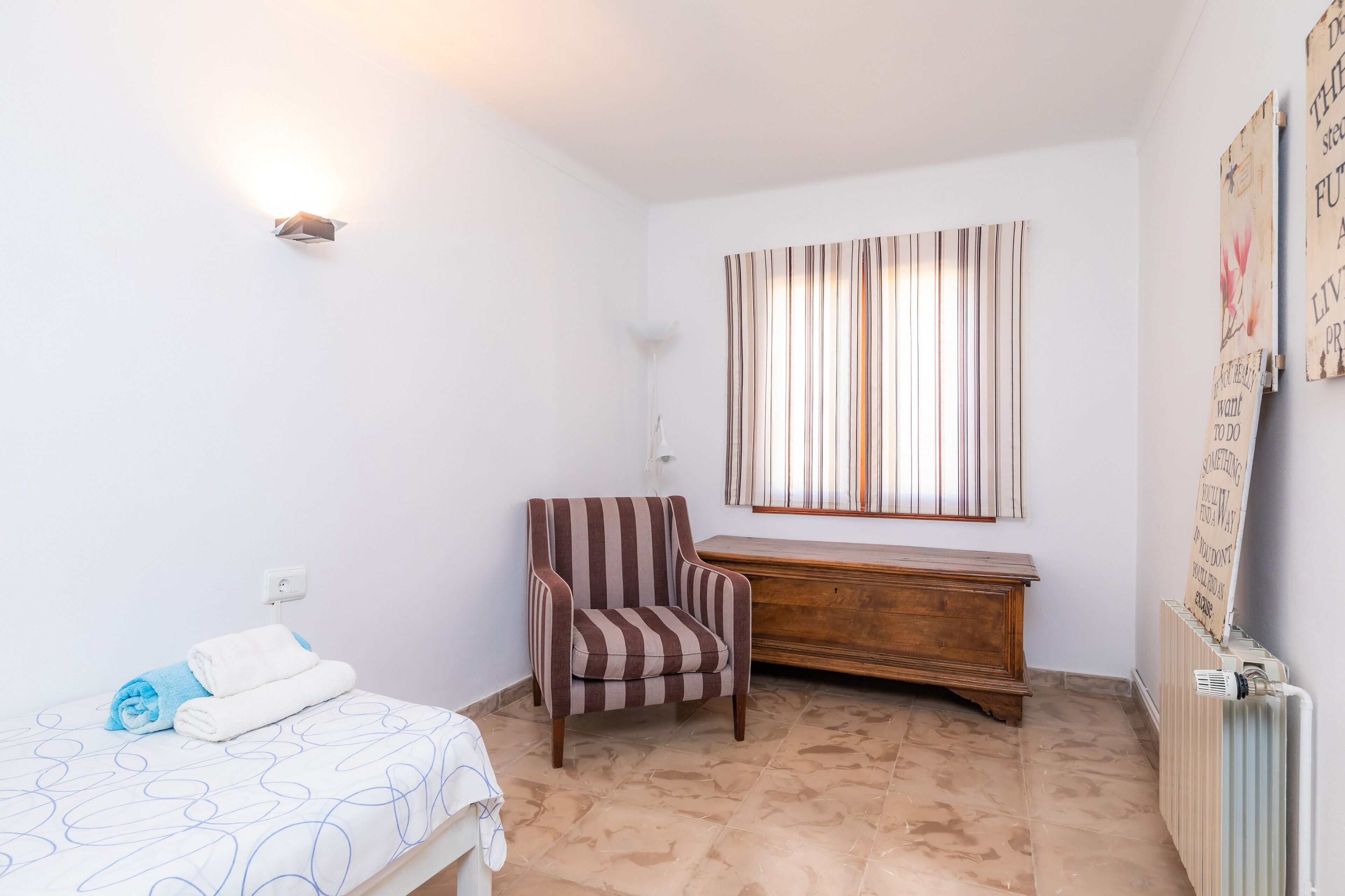 A bedroom with a bed, a striped armchair, a wooden dresser, a radiator, and a window with curtains.