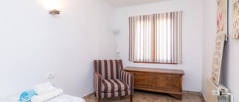 A bedroom with a bed, a striped armchair, a wooden dresser, a radiator, and a window with curtains.