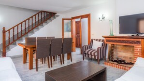Een woonkamer met een houten trap, een eethoek met stoelen, een bank, een open haard en een televisie.