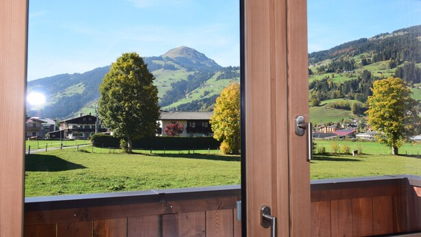 A view from a wooden-framed window overlooking a green landscape with trees, a mountain, and buildings.