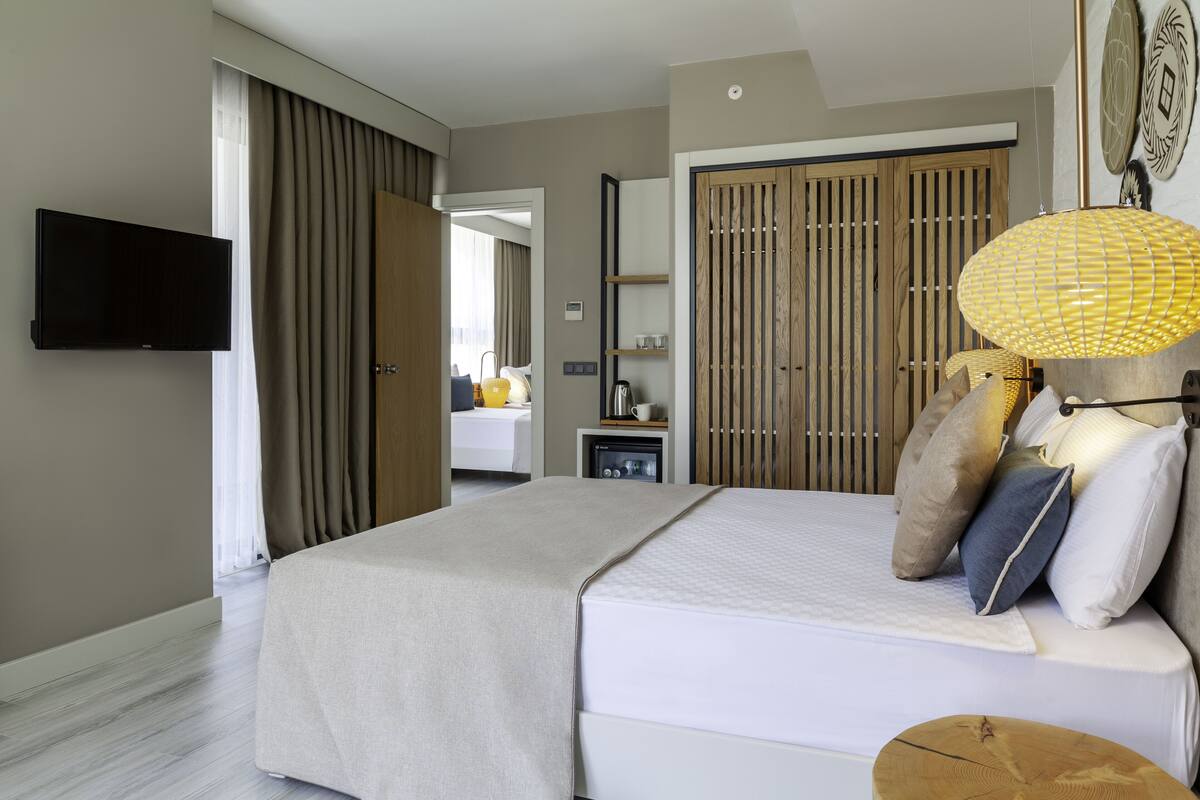Una habitación de hotel moderna con una cama grande, un televisor, un armario de madera y una mesita con una lámpara.