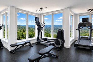 Un gimnasio con equipo de ejercicio, ventanas amplias y vista a un bosque.