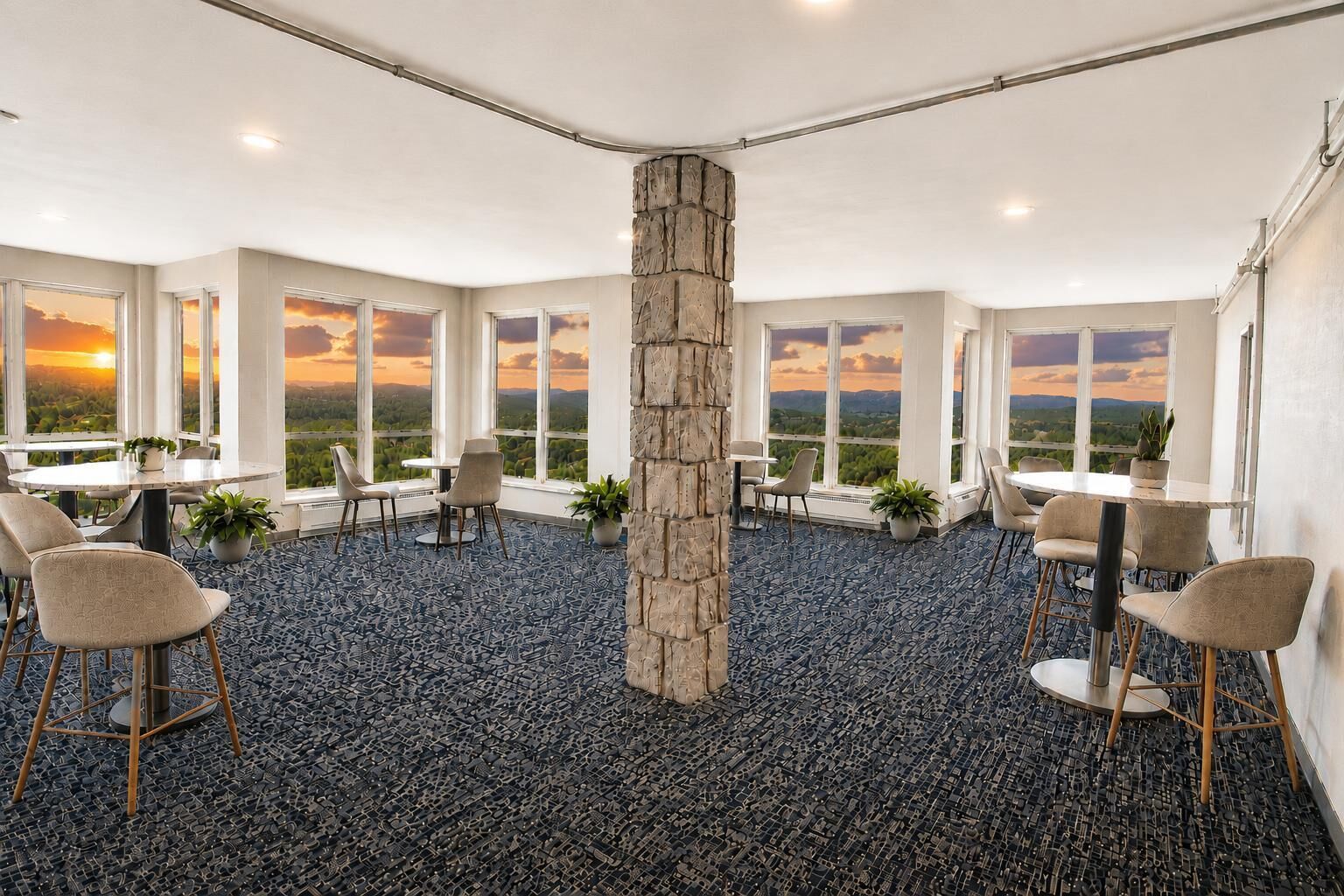 A spacious room with multiple tables and chairs, large windows with a view of the sunset, and a stone pillar in the center.