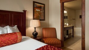 A hotel room with a large bed, a chair, a lamp, a painting, and a bathroom visible through an open door.