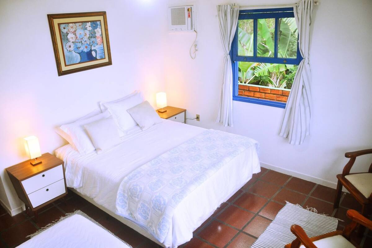 A neatly arranged bedroom with a bed, bedside tables, a nightstand, a window with curtains, a wall-mounted air conditioner, and a framed picture.