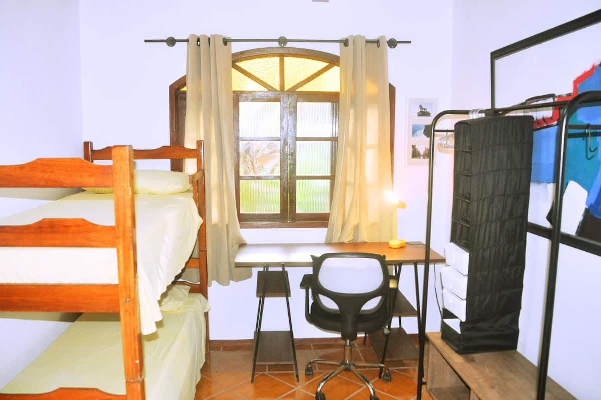 A dormitory room with bunk beds, a desk, a chair, and a window with curtains.