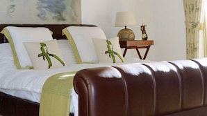 Ein ordentlich bezogenes Bett mit weißen Leinen und grünen Zierkissen, ein Nachttisch aus Holz mit einer Lampe sowie ein großes Bild an der Wand.
