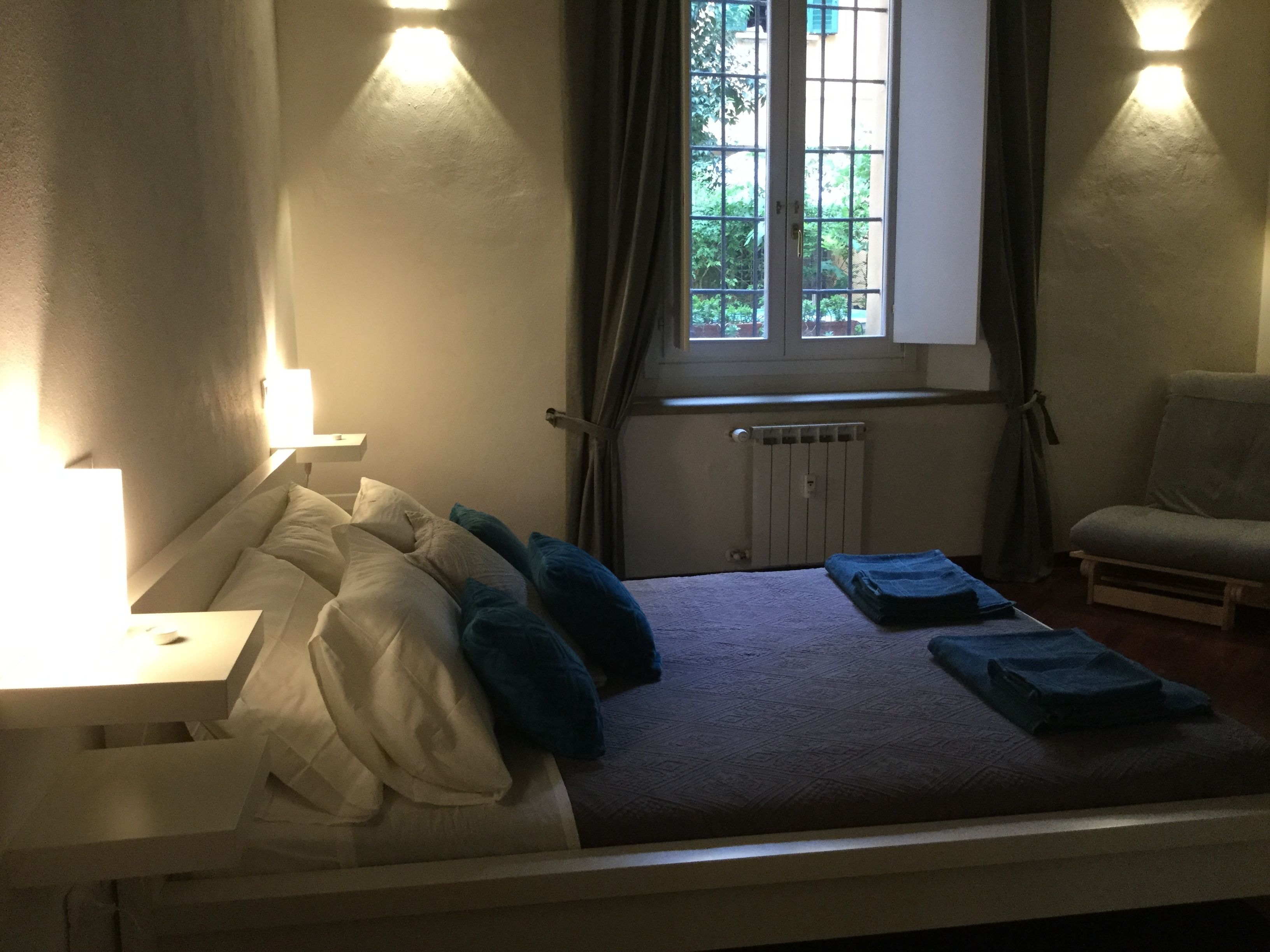 Un dormitorio con cama, almohadas, una mesita de noche, una silla, una ventana y un radiador.