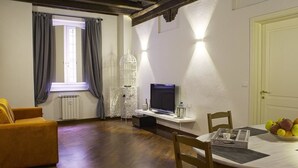 Ein gemütliches Wohnzimmer mit Sofa, Esstisch mit Stühlen, Fernseher und einem Fenster mit Vorhängen.