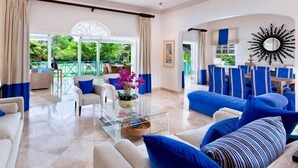A spacious living area with a glass coffee table, blue and white sofas, and a view of a lush garden through large windows. - Villa Moon Dance (Paynes Bay)