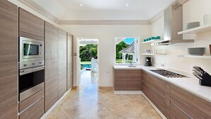 A modern kitchen with wooden cabinets, a built-in oven, microwave, and stove. There is a window with a view of greenery and a white ceiling. - Villa Moon Dance (Paynes Bay)
