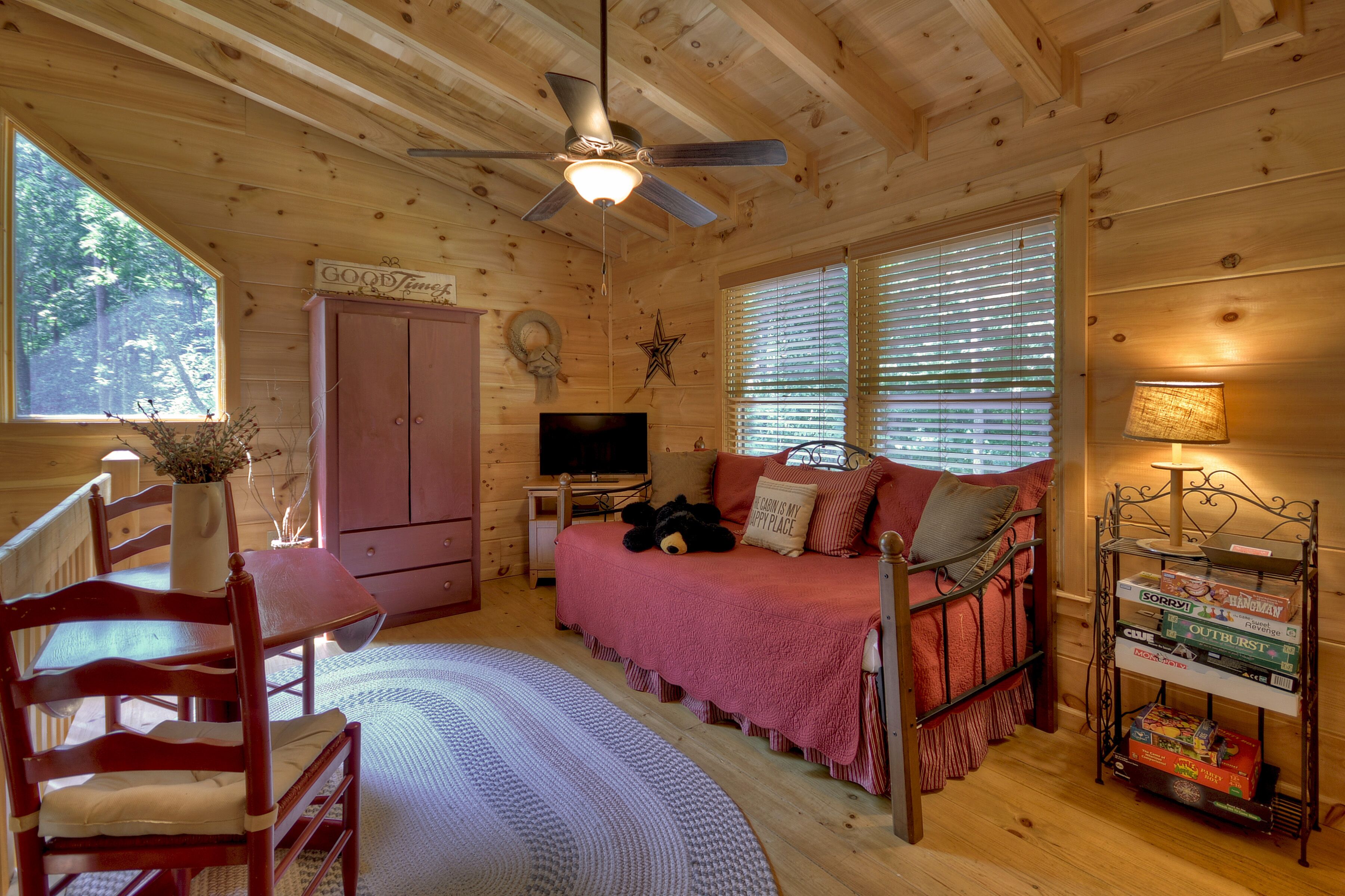 A cozy cabin interior with a wooden ceiling, a ceiling fan, a TV, a sofa with a blanket, a dining table with chairs, and a bookshelf with books.
