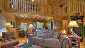 Interior acolhedor de uma cabana, com decoração em madeira, um sofá de couro, área de refeições e lareira.