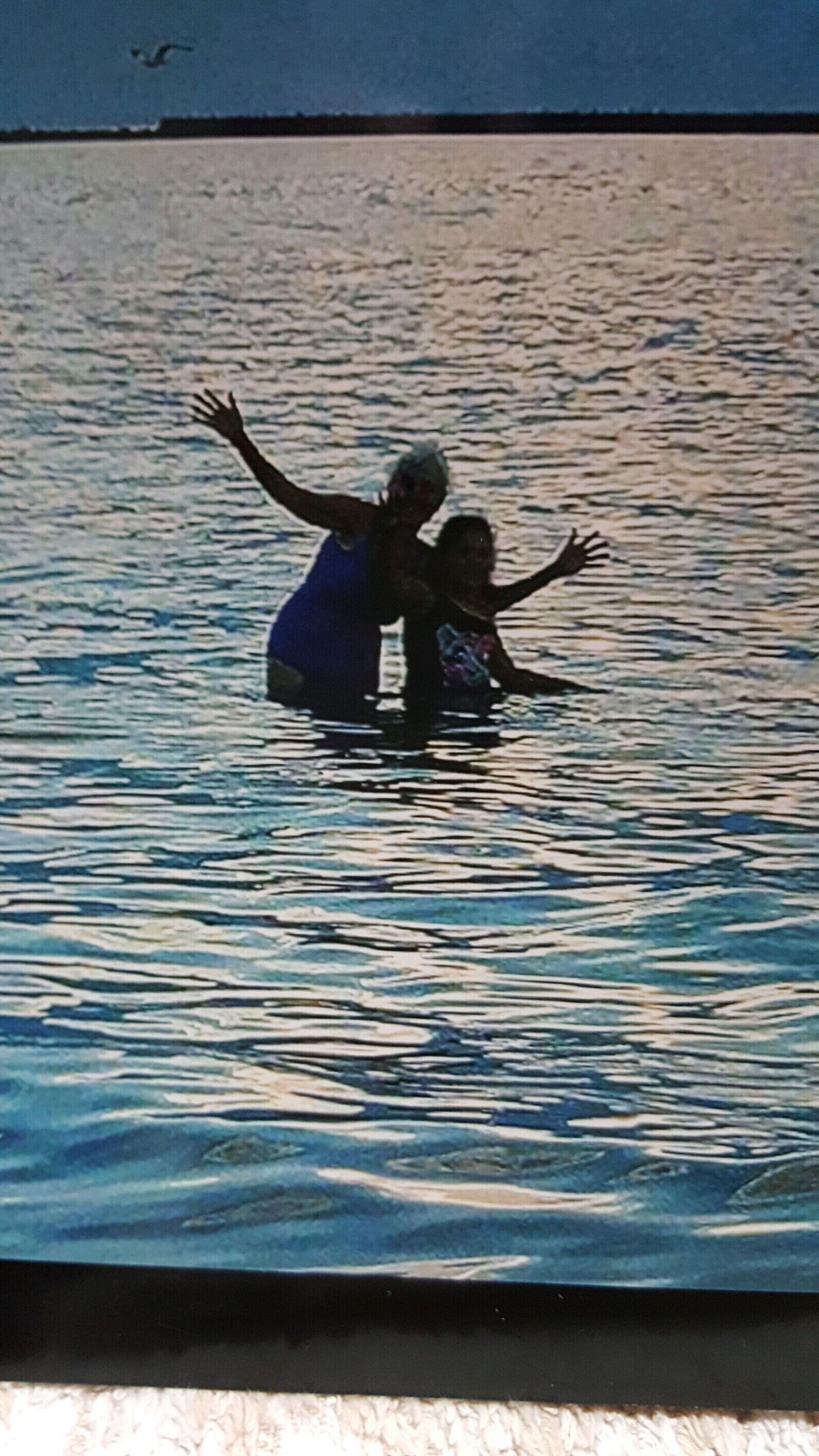 A person in a blue swimsuit standing in shallow water with arms raised.