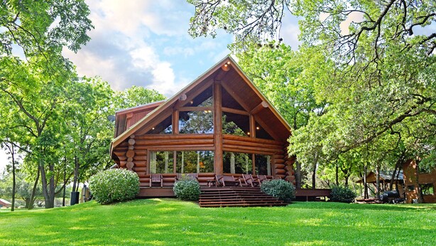 A log cabin with a porch and multiple windows, surrounded by lush greenery. - Lakefront Cabin w/ Loft • Sleeps 12 • Amenities (Burnet)