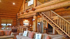 A log cabin interior with a wooden staircase, a dining area with a table and chairs, and a cozy living room with a sofa and cushions. - Lakefront Cabin w/ Loft • Sleeps 12 • Amenities (Burnet)