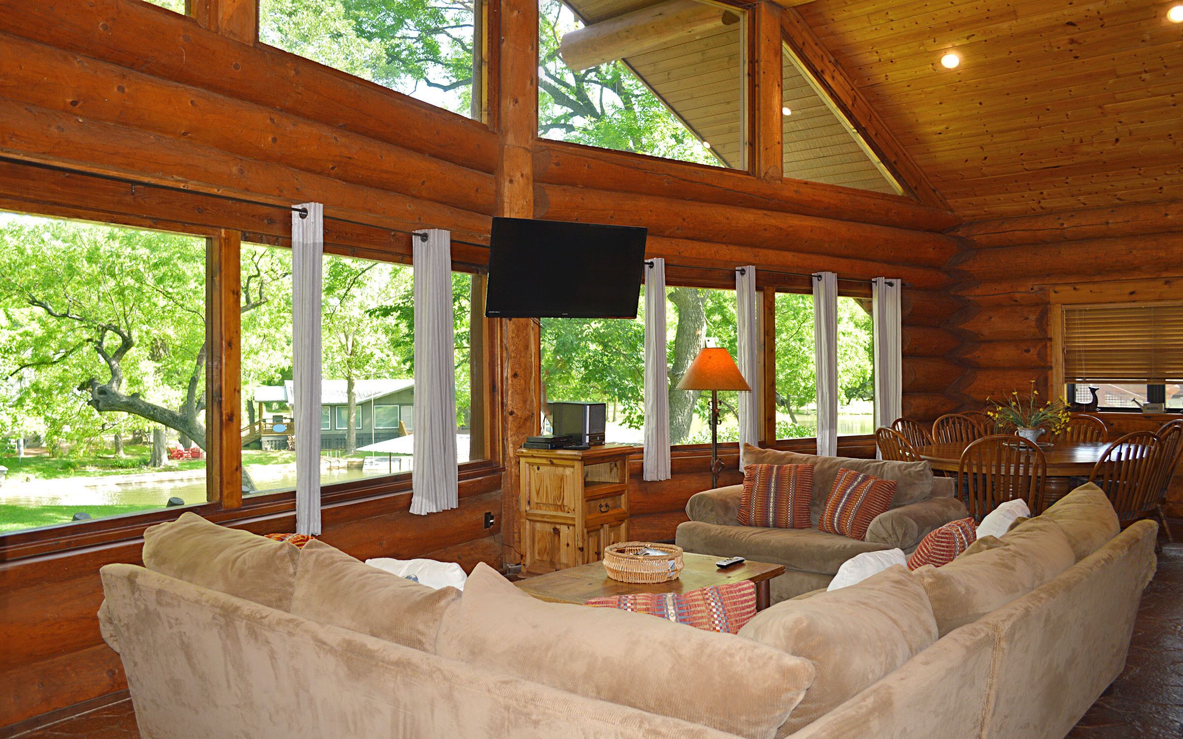 A log cabin interior with a large window, a flat-screen TV, a dining area, and a sofa with cushions.