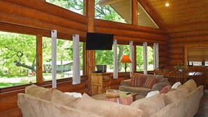 A log cabin interior with a large window, a flat-screen TV, a dining area, and a sofa with cushions. - Lakefront Cabin w/ Loft • Sleeps 12 • Amenities (Burnet)