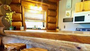 A wooden bar area with three stools, a wooden counter, and a kitchen area with a microwave and cabinets. - Lakefront Cabin w/ Loft • Sleeps 12 • Amenities (Burnet)