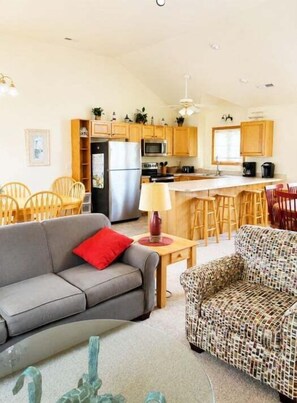 A modern kitchen with wooden cabinets, a refrigerator, and a microwave. There is a dining area with a table and chairs, and a living room with a sofa and a patterned armchair.