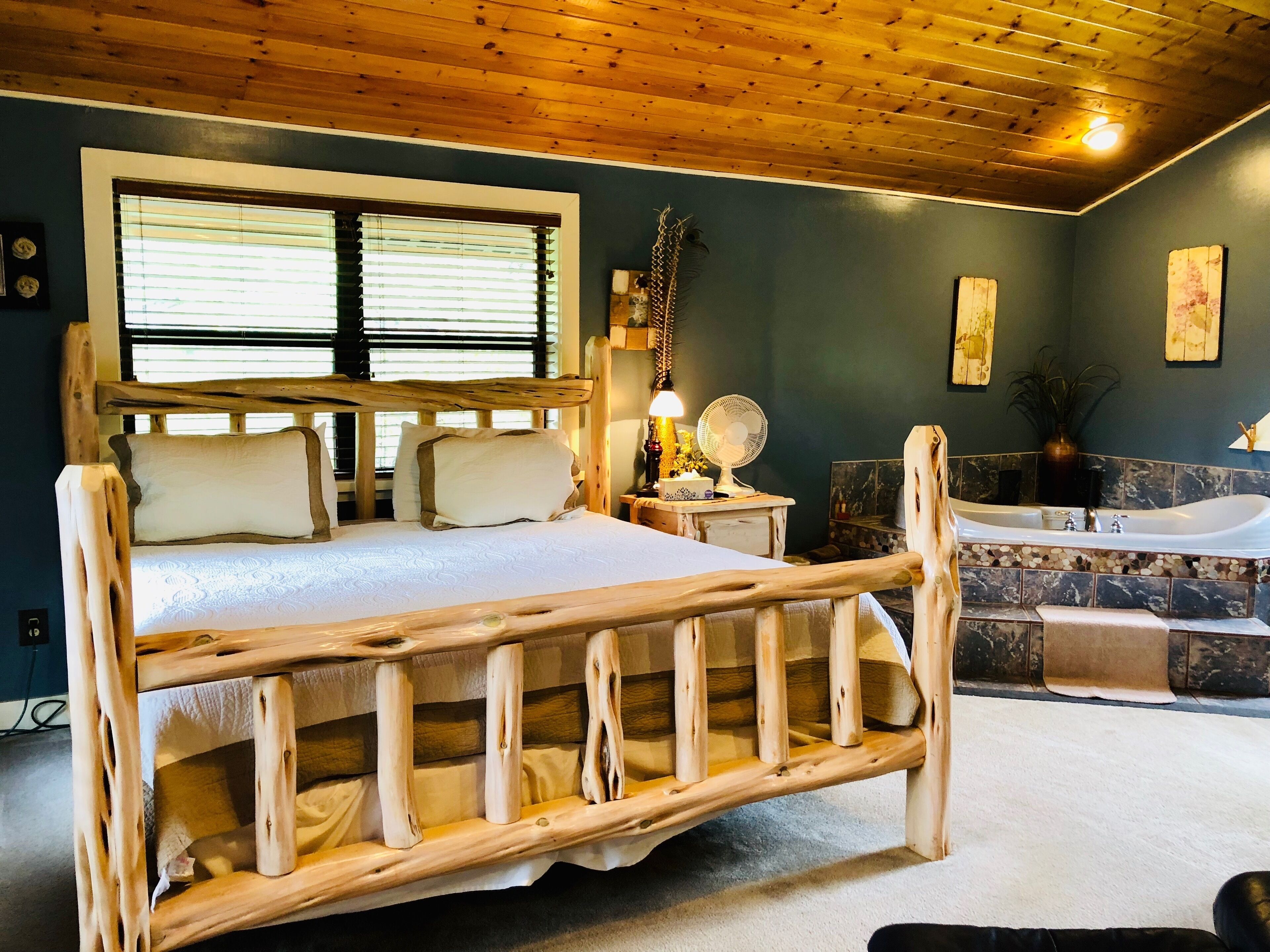 Un lit avec un cadre en bois, une grande fenêtre, une salle de bain avec une baignoire et un ventilateur posé sur une table de chevet.