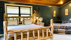 A wooden-framed bed with a large window, a bathroom with a bathtub, and a fan on a nightstand.