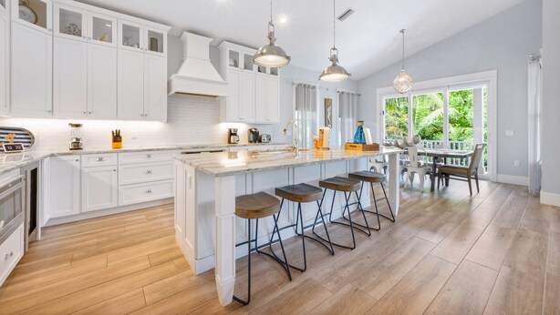 A modern kitchen with white cabinetry, a large island with bar stools, pendant lighting, and a view of greenery outside. - 4 Bed 4.5 Bath Family Friendly, 3 Blocks to Gulf (Anna Maria)