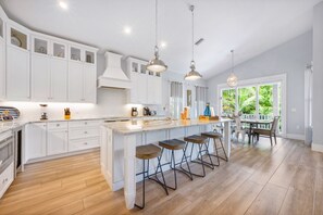 A modern kitchen with white cabinetry, a large island with bar stools, pendant lighting, and a view of greenery outside. - 4 Bed 4.5 Bath Family Friendly, 3 Blocks to Gulf (Anna Maria)