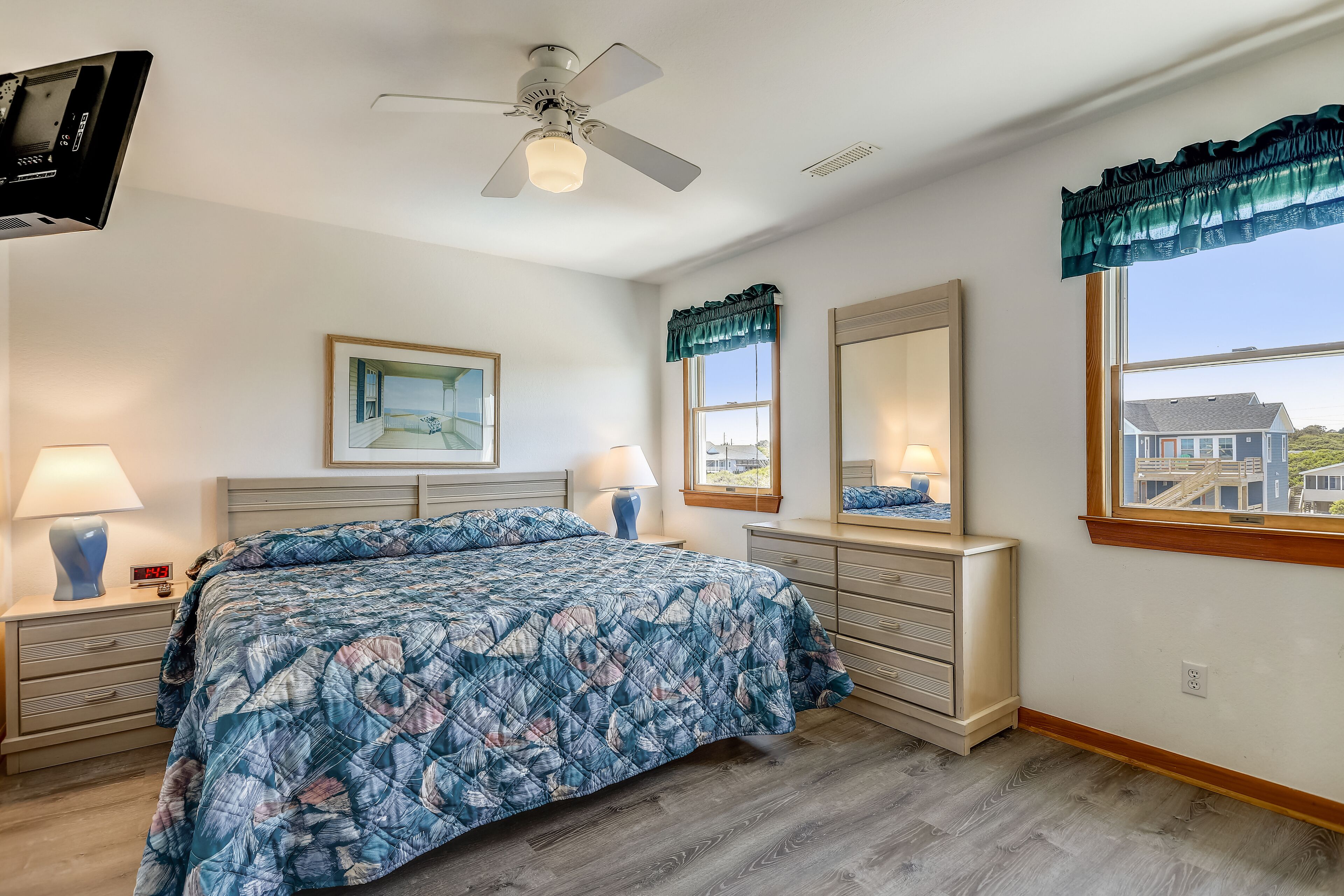 A bedroom with a bed, a nightstand, a dresser, a ceiling fan, a window with a view, and a framed picture on the wall.
