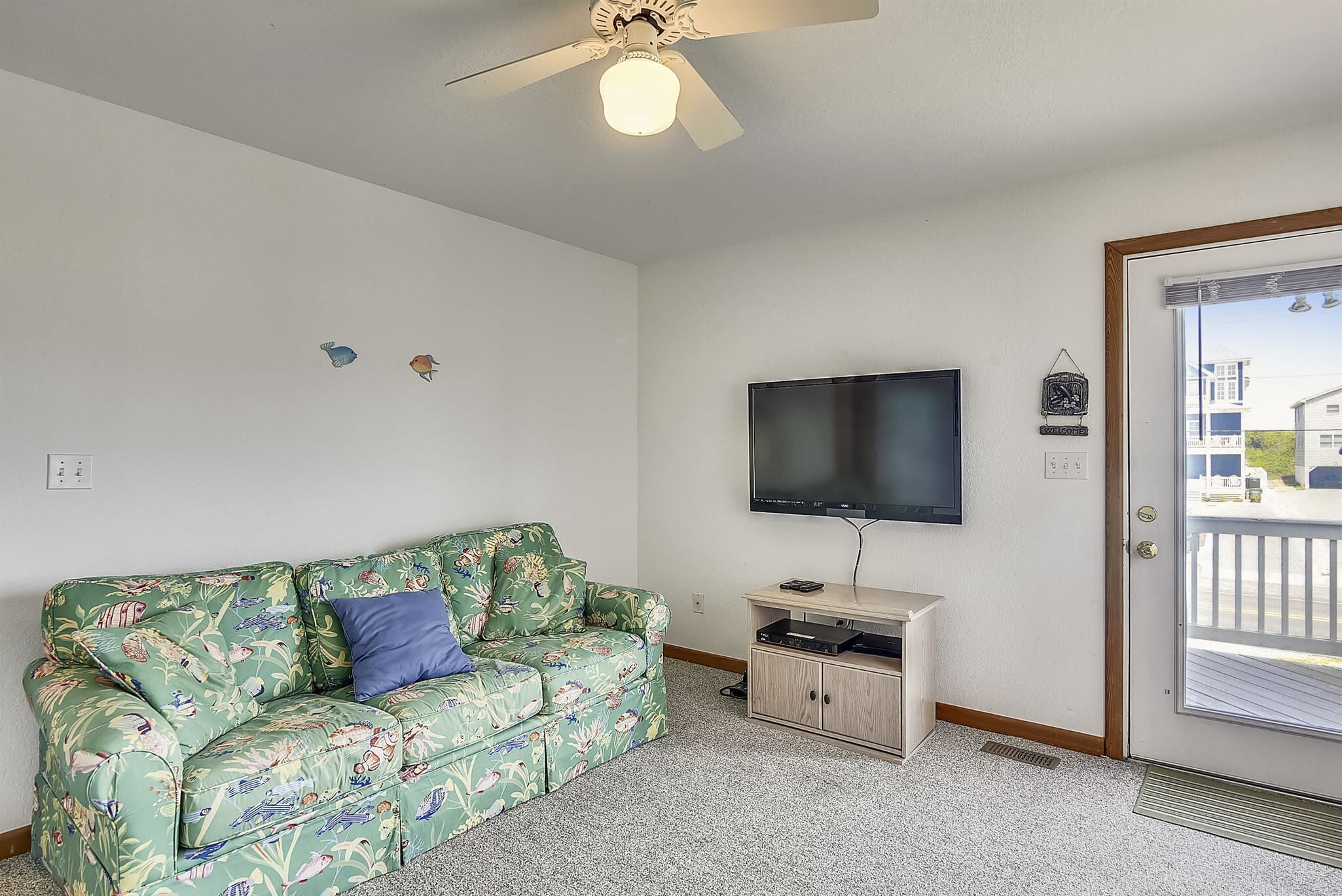 Una habitación con un sofá de flores verdes, un televisor de pantalla plana, un mueble de madera y una puerta con vista al exterior.