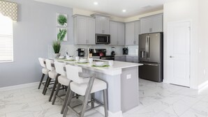A modern kitchen with grey cabinets, a stainless steel refrigerator, and a white island with bar stools. - Modern Luxury Villa with Everything (Kissimmee)