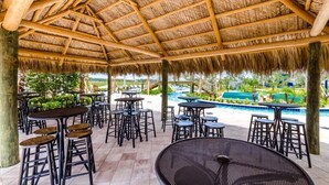 An outdoor patio area with thatched roof, tables, and chairs, overlooking a swimming pool and greenery. - Modern Luxury Villa with Everything (Kissimmee)