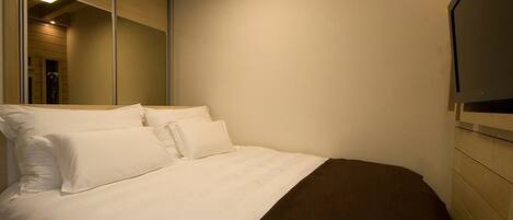 Een slaapkamer met een bed, kussens, een grote spiegel en een televisie aan de muur.