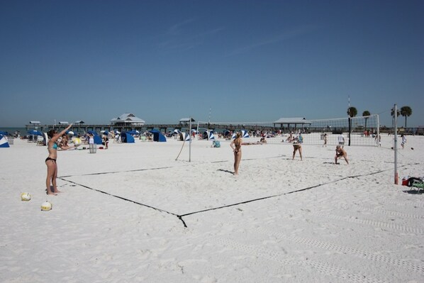 A beach volleyball court with players and spectators, a pier in the background, and beach umbrellas.