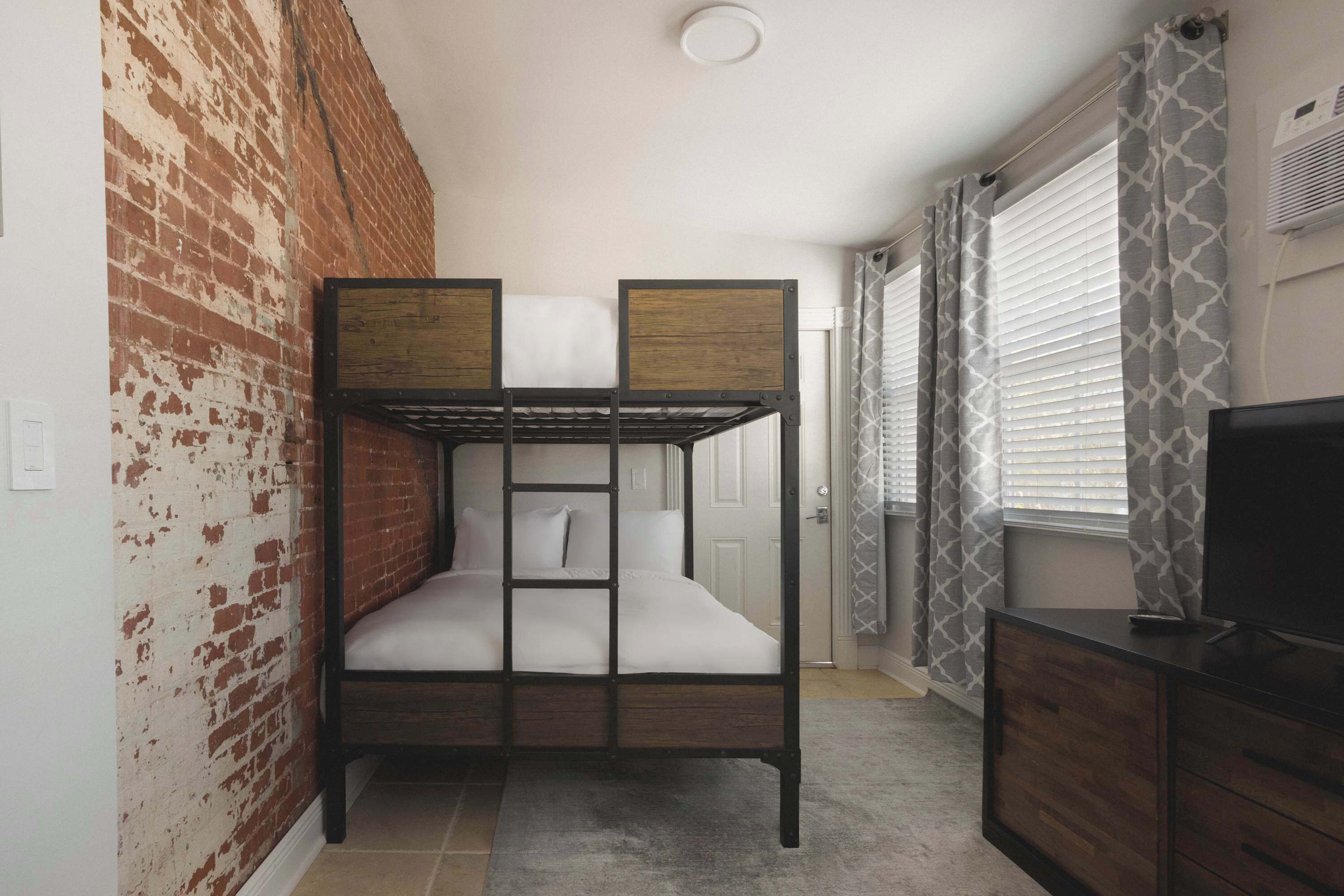 Een stapelbed met houten frame en wit beddengoed, een bakstenen muur, een raam met zonwering en een televisie op een houten dressoir.