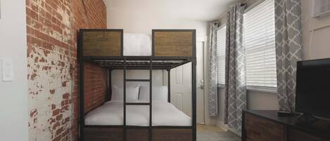 Een stapelbed met houten frame en wit beddengoed, een bakstenen muur, een raam met zonwering en een televisie op een houten dressoir.