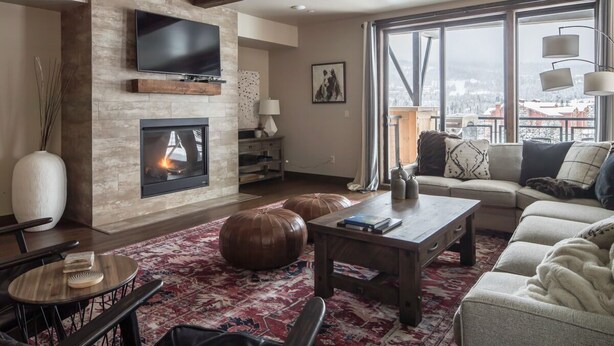 A living room with a fireplace, a television mounted on the wall, a sofa, a coffee table, and a view through large windows. - Easy Shuttle Access | Resort Amenities (Big Sky)