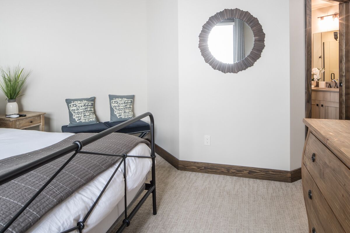 Quarto com uma cama de solteiro, mesa de cabeceira com uma planta, duas almofadas decorativas, um espelho redondo e um penteado de madeira.