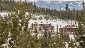A lodge with multiple floors, surrounded by snow-covered trees and mountains in the background. - Easy Shuttle Access | Resort Amenities (Big Sky)