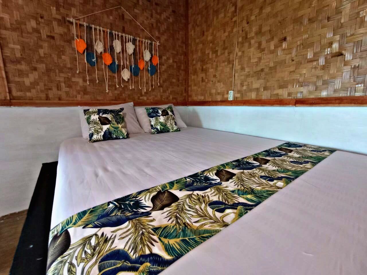 A bed with a tropical leaf-patterned bedspread, a hanging decorative item above the bed, and a woven wall.
