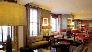 A spacious living room with a sofa, armchairs, and a coffee table. There are windows with curtains, a fireplace, and a dining area with a table and chairs. - Ardington Hotel (Worthing)