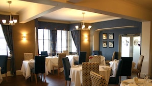 A restaurant with multiple tables and chairs, wooden flooring, and wall-mounted lighting. - Ardington Hotel (Worthing)