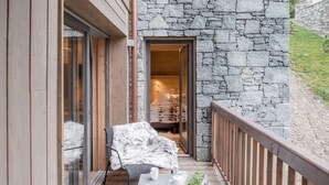 A balcony with a stone wall, a wooden deck, a chair, a table with a book, a teapot, and a plant. - Mammoth Lodge Apartment T3 Cabin D09 (Courchevel)
