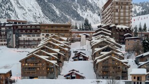Un complejo turístico en la montaña cubierto de nieve, con edificios de varios pisos, incluyendo un hotel destacado con balcones y un telesilla visible al fondo.