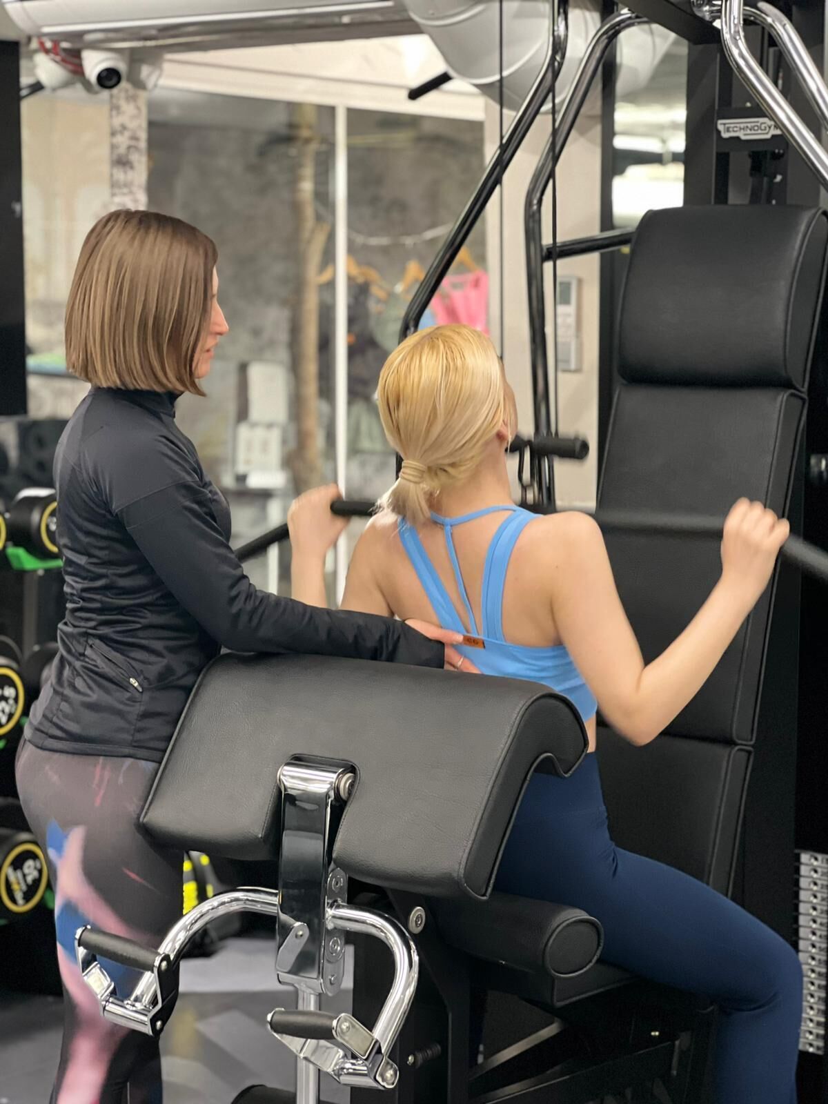 A gym with exercise equipment, a seated woman in workout attire, and a standing trainer.