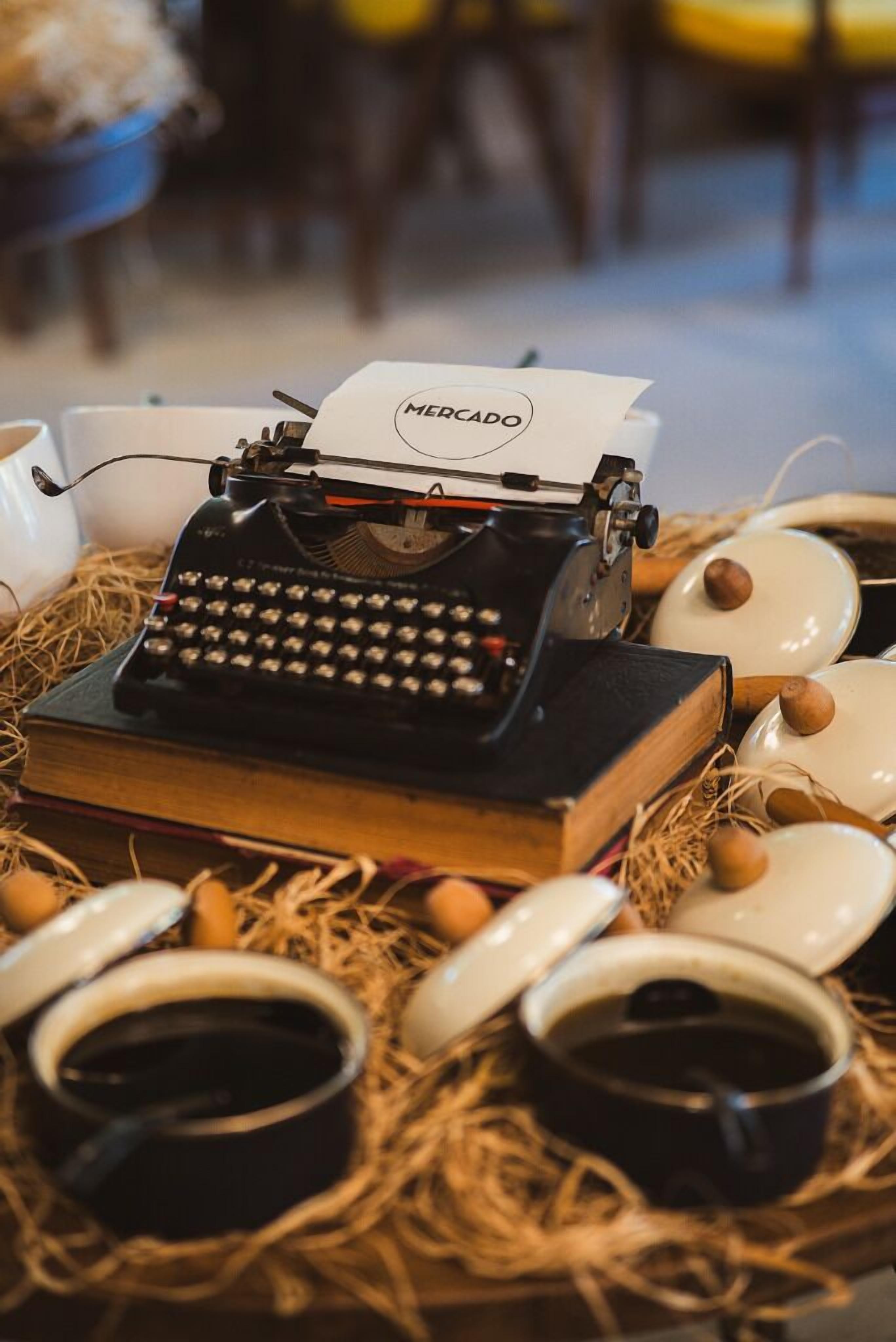 Uma máquina de escrever antiga com papel rotulado 'MERCADO' em cima, rodeada por xícaras de chá brancas e xícaras de café pretas sobre um tapete de palha.