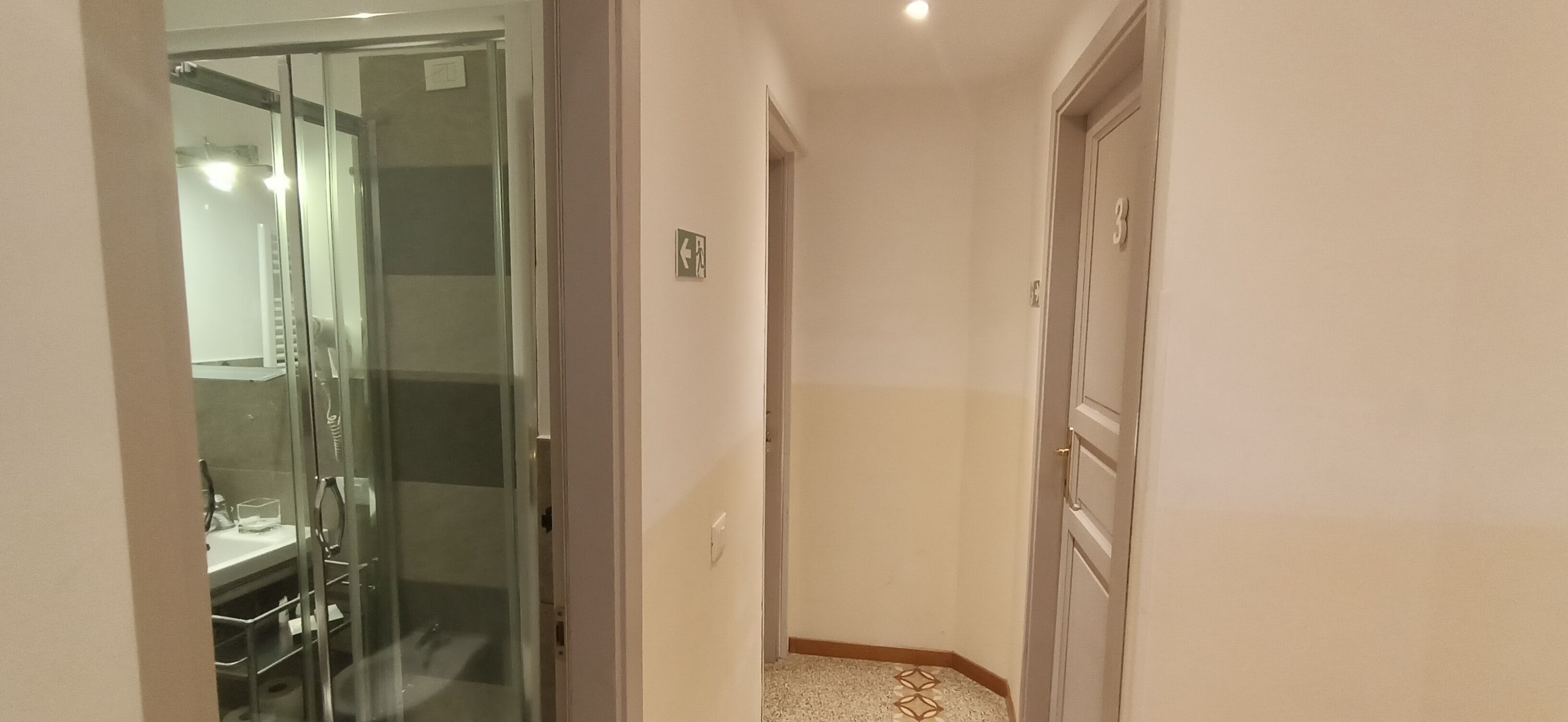 A narrow hallway with a glass door leading to a bathroom, a door with a number, and a small sign with an arrow.