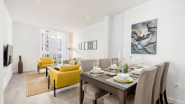 Un espace repas moderne avec une table en bois dressée pour quatre personnes, entourée de chaises rembourrées. Un canapé jaune et un téléviseur à écran plat sont visibles à l’arrière-plan.