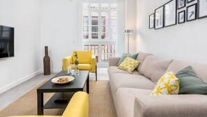 Un salon moderne avec un canapé, un fauteuil jaune et une table basse. Un téléviseur est fixé au mur et une fenêtre offre une vue sur les bâtiments.