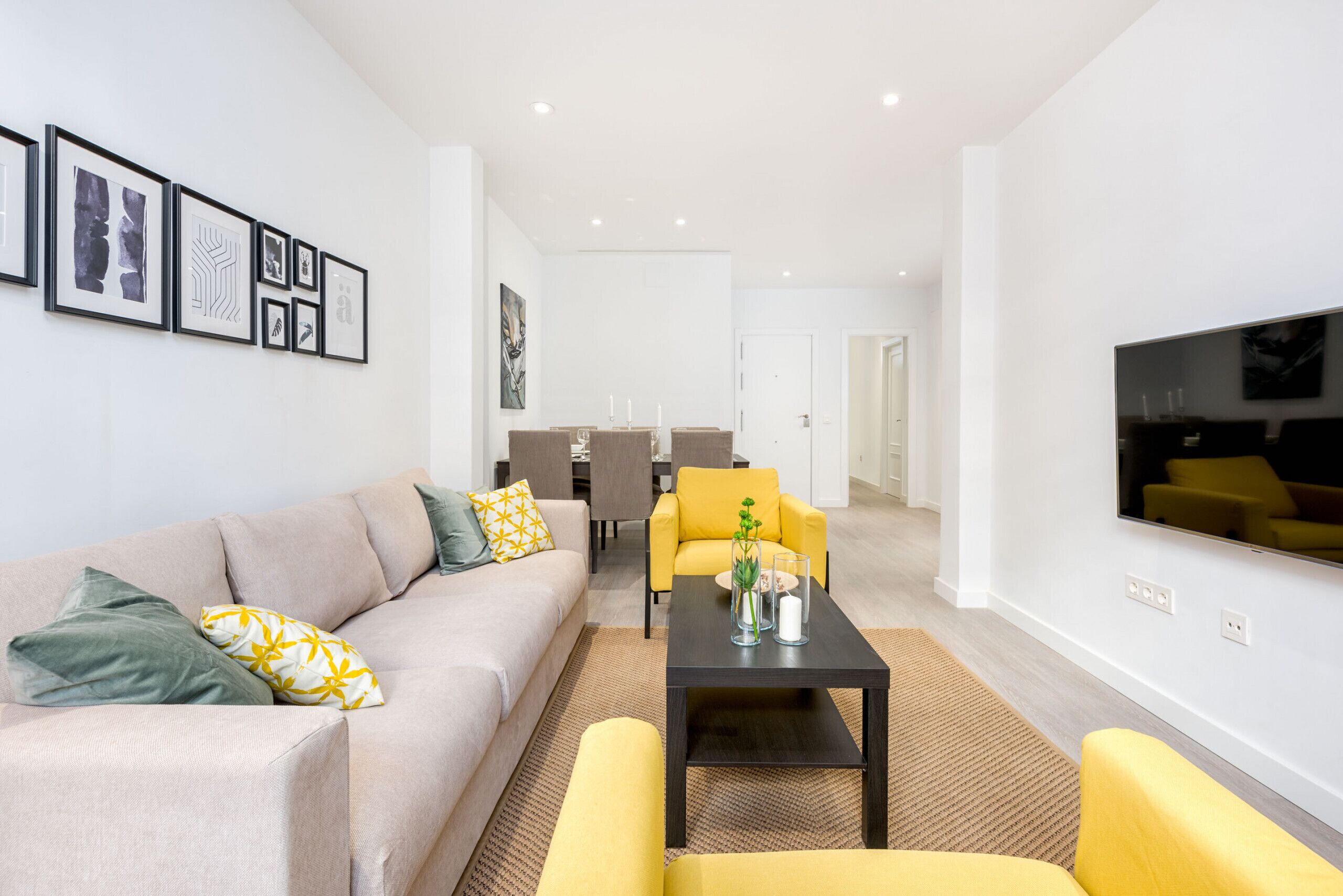A modern living room with a sofa, yellow armchairs, a coffee table, and a flat-screen TV mounted on the wall.
